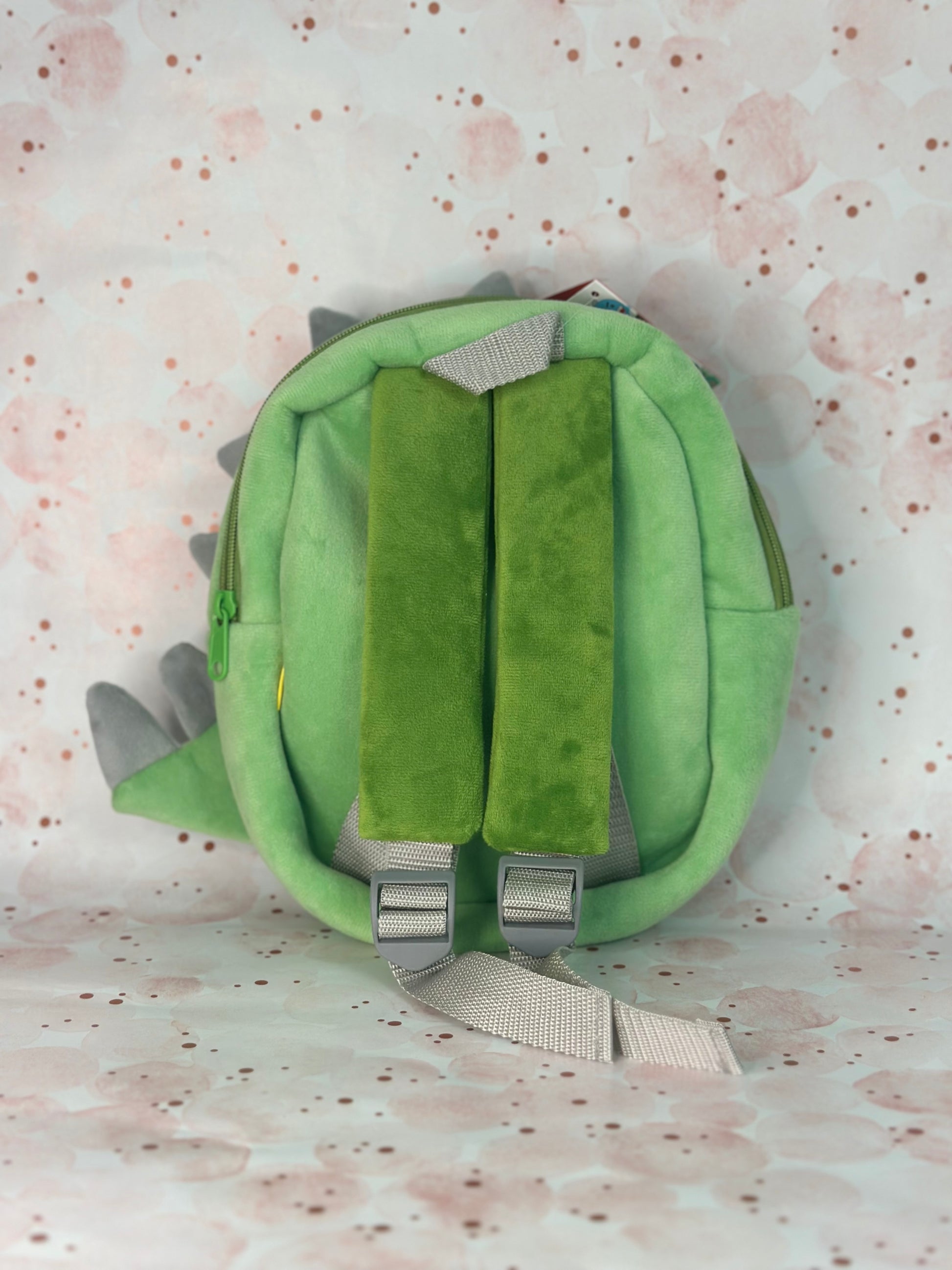 Green dinosaur-shaped backpack on a light pink background with subtle patterns